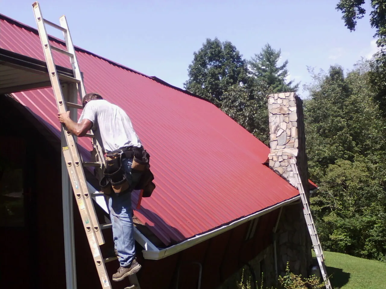 Roofer on a ladder beside a red metal roof and stone chimney — Soffit & Fascia Repair by Roofing Brothers in Matteson